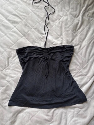Svart halterneck tubtopp med snörning - Snygg svart tubtopp med halterneckband och snörning framtill. Toppen har en lätt utsvängd passform och fina veckdetaljer upptill.
