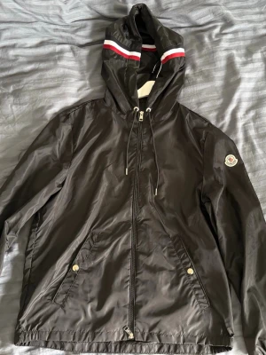 Svart vindjacka från Moncler - Snygg svart vindjacka från Moncler med huva och dragkedja framtill. Jackan har vita och röda ränder på huvan, två fickor med tryckknappar och Moncler-logga på ärmen. Tillverkad i ett glansigt, tunt material som står emot vind. Storlek L / M funkar 