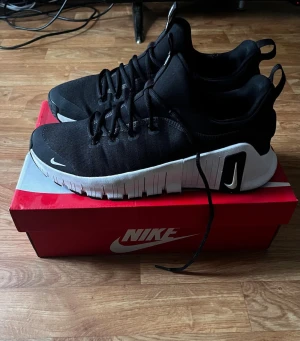 Svarta Nike Free sneakers - Snygga svarta Nike Free sneakers med vit sula och diskreta vita loggor. Ovandelen är i mesh-liknande material för bra andningsförmåga och skon har en sportig, modern look med dragflik bak och svarta skosnören. Perfekta för dig som gillar stilrena och bekväma sneakers.