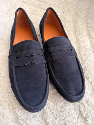 Mörkblå mocka loafers (oanvända) - Säljer ett par stilrena mörkblå loafers (helt oanvända) i mocka med klassisk penny strap och orange innersula. Skorna har en mjuk och följsam form med diskreta sömmar och passar perfekt till en avslappnad men ändå uppklädd look.