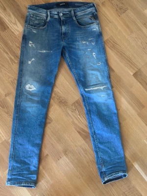 Blå slitna replay jeans  - Säljer ett par jeans från Replay med slitningar och distressed detaljer framtill. Jeansen har klassisk femficksdesign, normal midja och är tillverkade i denim med stretch för en bekväm passform.