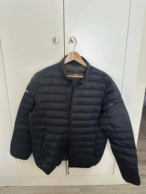 Svart pufferjacka från Elevate - Snygg svart pufferjacka från Elevate med quiltad design och dragkedja framtill. Jackan har två sidofickor med dragkedja och en extra ficka på ärmen. Perfekt för kalla vinterdagar och har en stilren look som passar till det mesta.