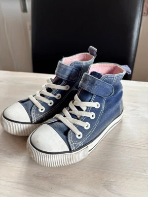 Blå höga sneakers med vita detaljer - Snygga blå sneakers i canvas med vita gummisulor och tåhättor. Skorna har vita snören, dekorativa sömmar och kardborreband upptill. Insidan är ljusrosa för en extra touch. Perfekta för en avslappnad och trendig stil.