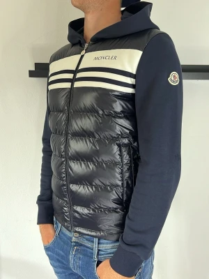 Moncler cardigan - Snygg marinblå hoodie från Moncler med vadderad, glansig framsida och vita ränder över bröstet. Dragkedja framtill, huva och Moncler-logga på ärmen. Ärmar och baksida i mjuk bomull, framsidan i blankt material. Perfekt för en sportig och trendig look.