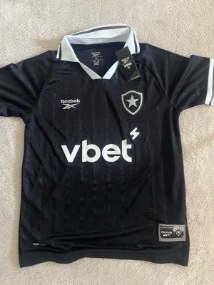 Svart fotbollströja från Reebok - Snygg svart fotbollströja från Reebok med vita detaljer och klubbmärke på bröstet. Tröjan har vit krage, korta ärmar med vita muddar och tryckt sponsorlogga framtill. Perfekt för dig som gillar sportig stil. Passar S/M