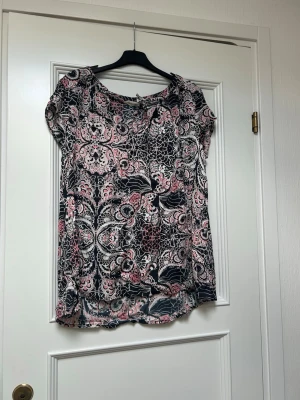 Mönstrad blus i rosa och svart - Säljer en luftig blus med korta ärmar och vringad hals. Blusen har ett detaljerat mönster i rosa, svart och vitt och är tillverkad i ett tunt, mjukt material som känns skönt mot huden. Oddmolly storlek 3