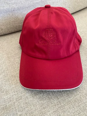 Röd keps från Loro Piana - Snygg röd keps från Loro Piana med broderad logga framtill. Kepsen har böjd skärm med vit kant och insidan har rutigt tyg samt brunt svettband. Justerbar baktill för perfekt passform. Perfekt accessoar för en sportig och stilren look.