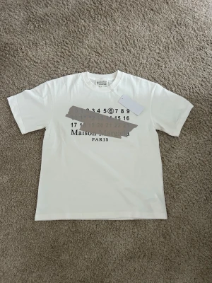 Vit Maison Margiela t-shirt med tryck - Vit t-shirt från Maison Margiela med ikoniskt siffertryck och grå tejpdetalj på bröstet. Klassisk passform med rund hals och korta ärmar. Perfekt för dig som gillar stilrena och unika plagg med designer-touch.