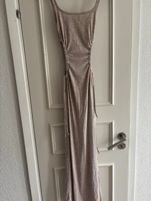 Beige långklänning med snörning - Snygg beige långklänning med smala axelband och snörning i sidorna som ger en smickrande passform. Klänningen har en rak siluett och är tillverkad i ett mjukt, stretchigt material som känns bekvämt mot huden.
