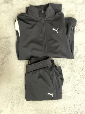Svart träningsset från Puma (13-14år) - Snyggt svart träningsset från Puma med jacka och byxor. Jackan har dragkedja och vit detalj på axeln, båda plaggen har Puma-logga framtill. Perfekt för träning eller chill. Materialet är mjukt och följsamt för bästa komfort.
