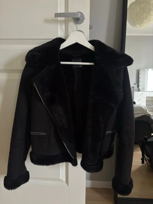 Svart teddyjacka med dragkedja - En svart teddyjacka med mjukt foder och bred krage. Jackan har dragkedja framtill, två fickor med dragkedjor och detaljer i fuskpäls längs krage, ärmslut och nederkant. Perfekt för kalla cozu dagar och riktigt snygg till jeans. Storlek är M men skulle nog säga det även passar som S. 