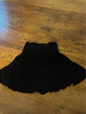 Svart kortkjol med volang - Supersöt svart kortkjol med volangkant och bred, smockad midja. Kjolen har en flowy känsla och passar perfekt till en trendig vardagslook. Enkel att matcha med både crop tops och stickade tröjor.