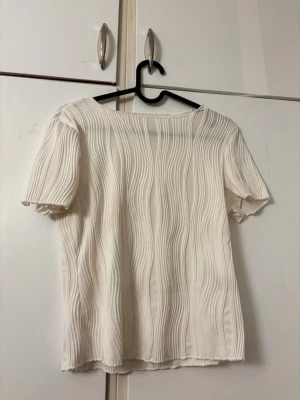 Vit ribbad topp med kort ärm - Snygg vit topp med korta ärmar och ribbad struktur. Toppen har en rak passform och är tillverkad i ett mjukt material som känns skönt mot huden. Perfekt att styla med jeans eller kjol för en fräsch look.