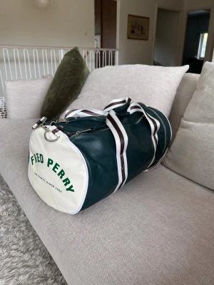 Fred Perry Barrel väska  - Snygg väska från Fred Perry i mörkgrönt och vitt läder. Väskan har rund form, vita detaljer och grönt tryck på sidan. Utrustad med randiga axelremmar och dragkedja upptill. Perfekt för dig som vill sticka ut med en sportig och retro look.