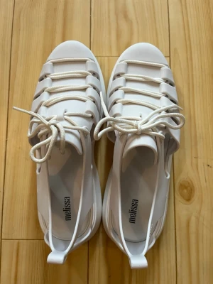 Vita sandaler med snörning från Melissa - Snygga vita sandaler från Melissa med cool snörning framtill och öppen design på sidorna. Tillverkade i ett mjukt, flexibelt plastmaterial som är lätt att rengöra. Perfekta för sommaren och ger en modern vibe till din outfit.