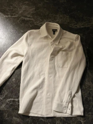 Vit overshirt med bröstficka - Stilren vit overshirt med klassisk krage och knappstängning framtill. Modellen har en bröstficka på vänster sida och långa ärmar med knapp vid ärmslut. Perfekt att bära som lager över t-shirt. Materialet ser ut att vara mjukt och något tjockare bomull.