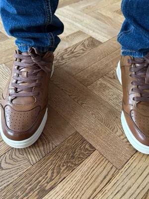Bruna Ralph Lauren skor - Snygga bruna sneakers i läder med vita sulor och klassisk snörning. Skorna har perforerade detaljer på tån och en stilren siluett som passar till jeans eller andra casual outfits. Perfekt för dig som gillar en clean och tidlös look.