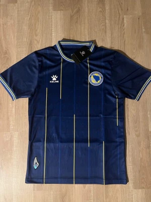 Blå fotbollströja Bosnien  - Snygg blå fotbollströja med Bosnien och Hercegovinas landslagsemblem på bröstet. Tröjan har gula och blå tunna ränder, ribbade muddar med gula och vita detaljer samt korta ärmar. Perfekt för match eller träning.
