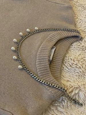 Silverfärgat halsband med pärlor - Snyggt halsband i silverfärgad rostfritt med kedja och små vita pärlor som hänger längs hela halsbandet. Perfekt för att ge en cool touch till din outfit. Passar till många olika stilar och är lätt att matcha med andra accessoarer.