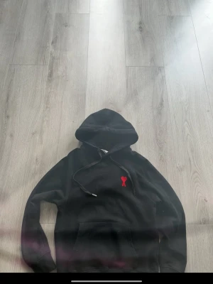 Svart hoodie med rött hjärta och A - Snygg svart hoodie med huva och känguruficka. På bröstet sitter en broderad röd detalj i form av ett hjärta och bokstaven A. Hoodien har dragsnören vid huvan och är tillverkad i mjukt material, perfekt för en chill och stilren look.