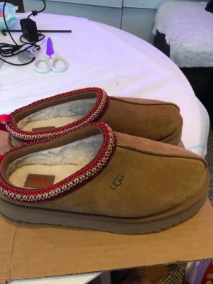 Bruna UGG tofflor med röd kant - Säljer ett par bruna UGG tofflor i mocka med mjukt vitt foder och en snygg röd och vit mönstrad kant runt öppningen. Perfekta för att hålla fötterna varma hemma. Klassisk UGG-logga på sidan och rejäl sula.
