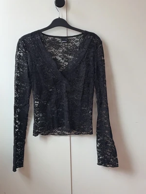 Svart spetsblus med v-ringning - Snygg svart blus i genombruten spets med v-ringad hals och långa ärmar. Perfekt för dig som vill ha en trendig och elegant topp med transparent känsla och fina spetsdetaljer. Passar till både jeans och kjol för en cool look.