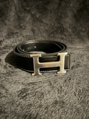 Svart skärp med silverfärgat H-spänne - Snyggt svart bälte i skinn med ett stort, silverfärgat H-format spänne. Bältet har vita sömmar längs kanterna som ger en stilren kontrast. Perfekt accessoar för att lyfta din outfit och ge en exklusiv touch.