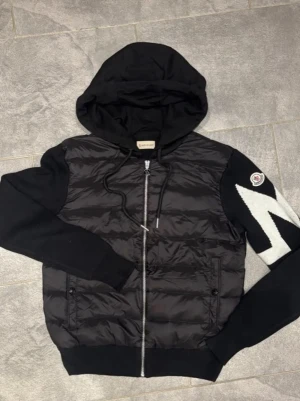 Svart Cardigan - Snygg svart hoodie med vadderad framsida och dragkedja. Ärmarna är i mjukt tyg, ena ärmen har ett vitt zigzag-mönster och en patch på axeln. Huva med dragsko och två fickor framtill. Perfekt för en streetwear-look.