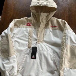 Vit och beige jacka från Supreme och Umbro - En vit och beige jacka från Supreme och Umbro med röda logotyper på bröstet och ryggen. Jackan har en halv dragkedja framtill och en huva.