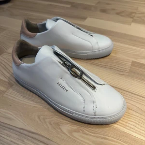 Axel Arigato skor - Säljer dessa vita Axel Arigato skor med rosa detalj i hälen. Väldigt fint skick. Storlek 38. Påsen på bilden medföljer