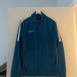 Nike  - En blå Nike-jacka med vita detaljer. Jackan har en dragkedja framtill och två fickor på sidorna. Den har en vit swoosh-logotyp på bröstet.