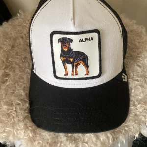 Keps - En The Farm keps med en text där det står ”Alpha”. Kepsen har också en bild på en hund. 