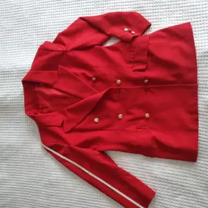 Trä in i kungadömet av hög elegans med denna vintage röda kappa som är här för att regera din garderob. Prydd med guldiga knappar på båda sidor är det inte bara en kappa; det är en majestätiskt modeuttalande. Fantastiskt form 