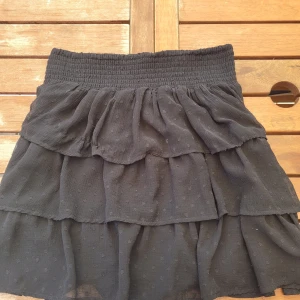 Svart kjol från Lindex - En svart kjol från Lindex med volanger och elastisk midja. Kjolen har ett diskret prickigt mönster och är tillverkad i ett lätt och luftigt material. Använd fåtal gånger.