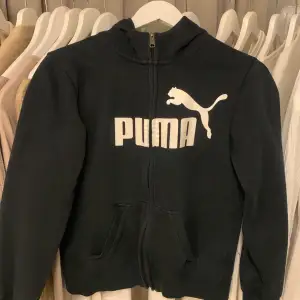 En svart jacka från Puma med dragkedja och långärmad design. Jackan har en vit Puma-logotyp på framsidan. sitter mer som strl S