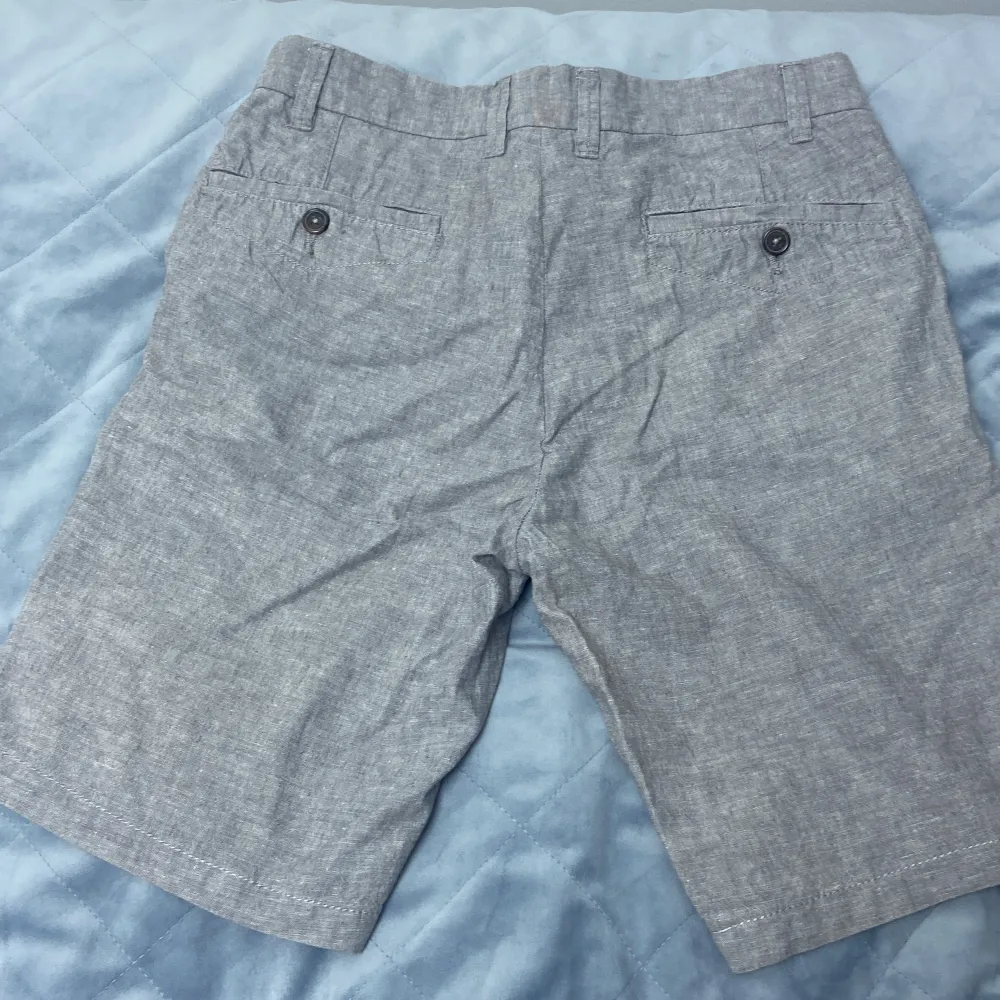 Ett par grå shorts från Dressmann i linneblandning. Shortsen har två framfickor och två bakfickor med knappar. De har en knapp och dragkedja framtill.. Shorts.