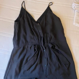 Svart långklänning med smala axelband - En elegant svart långklänning (dress med ben) med smala axelband och knappar framtill. Klänningen har en v-ringad hals och en åtsittande midja som ger en smickrande passform. 