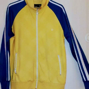 Sweatshirt gul/blå med vita ränder - En skön sweatshirt med dragkedja gjord av 50 % bomull och 50 % polyester. Funkar till vardags som till träning. Fickor med zipkedjor. Inte mycket använd då den är för liten för mig.  Mått: B:55 cm. Längd: 70 cm