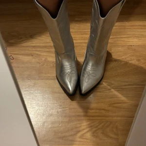 Stilrena silvriga cowboy boots med liten klack, storlek 37 👢 - Säljer stilrena silvriga cowboy boots i storlek 37 med liten klack. Dessa boots ger ett unikt och trendigt uttryck och är perfekta för att lyfta din outfit. Helt oanvända.