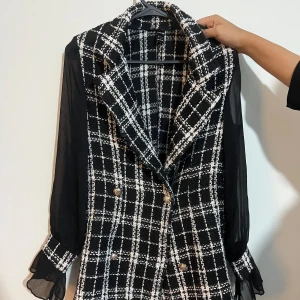 Svart och vit rutig kavaj med genomskinliga ärmar - En elegant svart och vit rutig kavaj med genomskinliga långa ärmar. Kavajen har en dubbelknäppt design med guldknappar och en klassisk krage. Ärmsluten har volangdetaljer. Köpt från shein men BRA MATERIAL OCH KVALITET. 