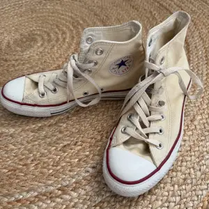 Ett par beige höga sneakers från Converse med snörning och vit sula. Skorna har den klassiska Converse-logotypen på sidan och en röd rand längs sulan.