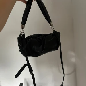 Svart handväska i skinn - En elegant svart handväska i skinn med en mjuk, rynkad design. Väskan har en kort handrem och en längre axelrem som kan justeras eller tas bort. Insidan är rymlig med flera fack för organisering.