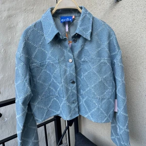Blå denim jacka från Crās - En blå jacka från Crās med ett rutigt mönster. Jackan har långa ärmar, en krage och knappar framtill. 