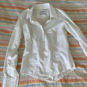Vit skjorta från The Shirt Factory - En elegant vit skjorta från The Shirt Factory, designad av Linnéa Braun. Skjortan är tillverkad av 100% bomull och har en klassisk krage samt knappar framtill. Den har långa ärmar med manschetter och en figursydd passform.