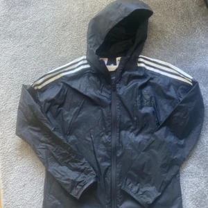 Svart vindjacka från Adidas - En svart vindjacka från Adidas med huva och dragkedja. Jackan har vita ränder på axlarna och ett stort nummer 96 på ryggen. Adidas-logotypen är tryckt på bröstet. knappt använd