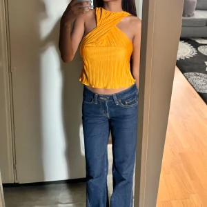 Gul/Orange Topp - Toppen har en unik design med korsade band över bröstet och en hög hals. Materialet är plisserat och lätt, vilket ger en elegant struktur och en bekväm passform. Perfekt för sommarens varma dagar eller för att lägga till en färgklick i din outfit.