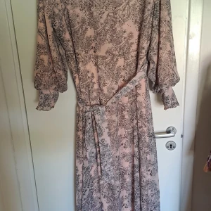 Blommig klänning från Alia - En vacker blommig klänning från Alia i storlek EU 52. Klänningen har en ljusrosa bas med ett detaljerat svart blommönster. Den har långa ärmar med volangdetaljer vid ärmsluten och en knytning i midjan. Klänningen har en dragkedja i ryggen.