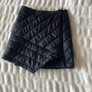 Svart termo kjol från Everest - En svart termokjol från Everest med diagonala sömmar och dragkedja på sidan. Kjolen är kort och har en elastisk midja för bättre passform.