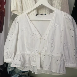 Vit blus med broderade detaljer - En vit blus med broderade detaljer och knappar framtill. Blusen har trekvartsärmar och en volangkant nedtill. Den är tillverkad i ett lätt och luftigt material, perfekt för varmare väder.