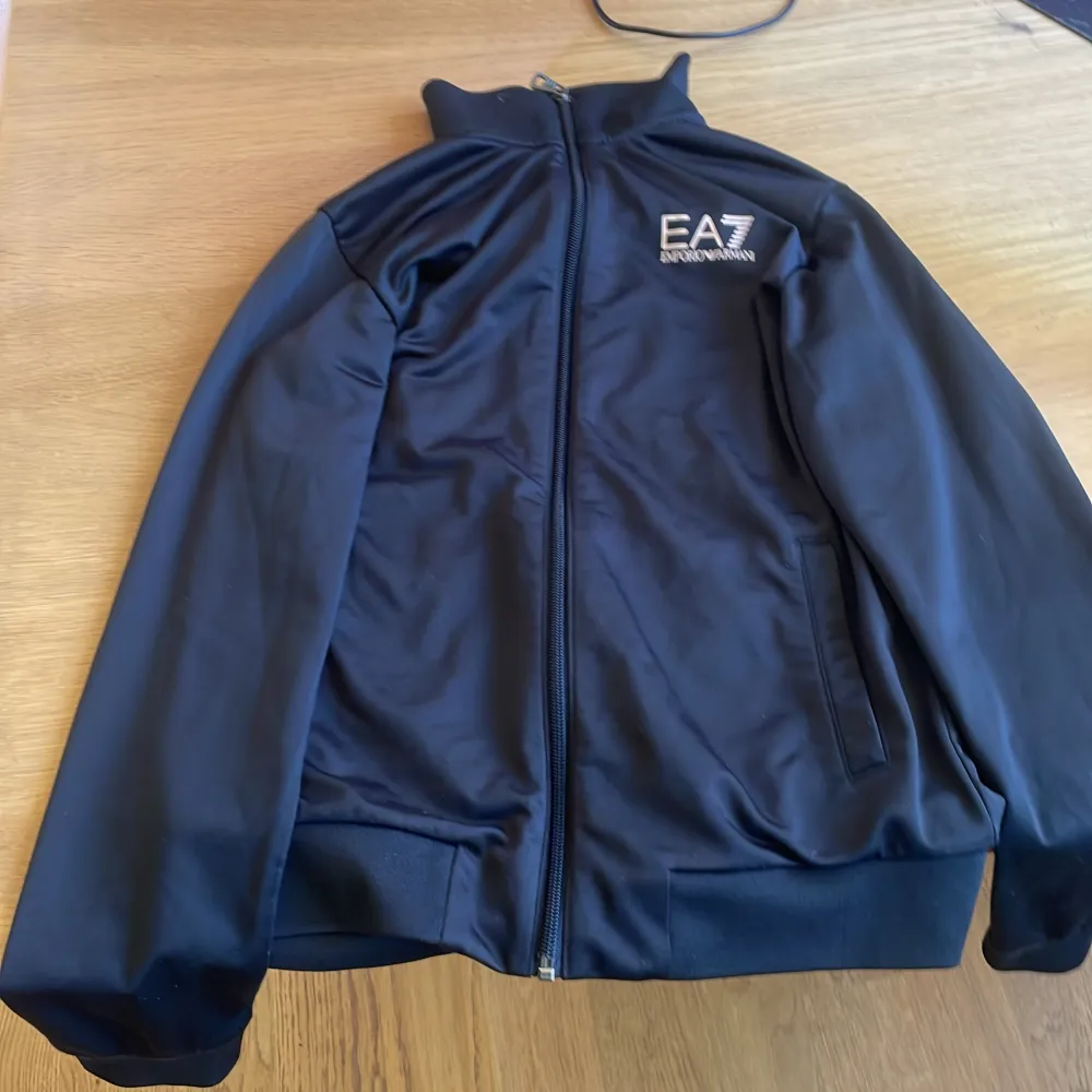 EA7 Emporio Armani-tröjan, storlek 14A (160 cm), är begagnad men har bara använts några få gånger, maximalt 10 gånger. Trots sin begränsade användning är den fortfarande i utmärkt skick och bibehåller sin höga kvalitet och stil. Matchande byxor finns. Tröjor & Koftor.